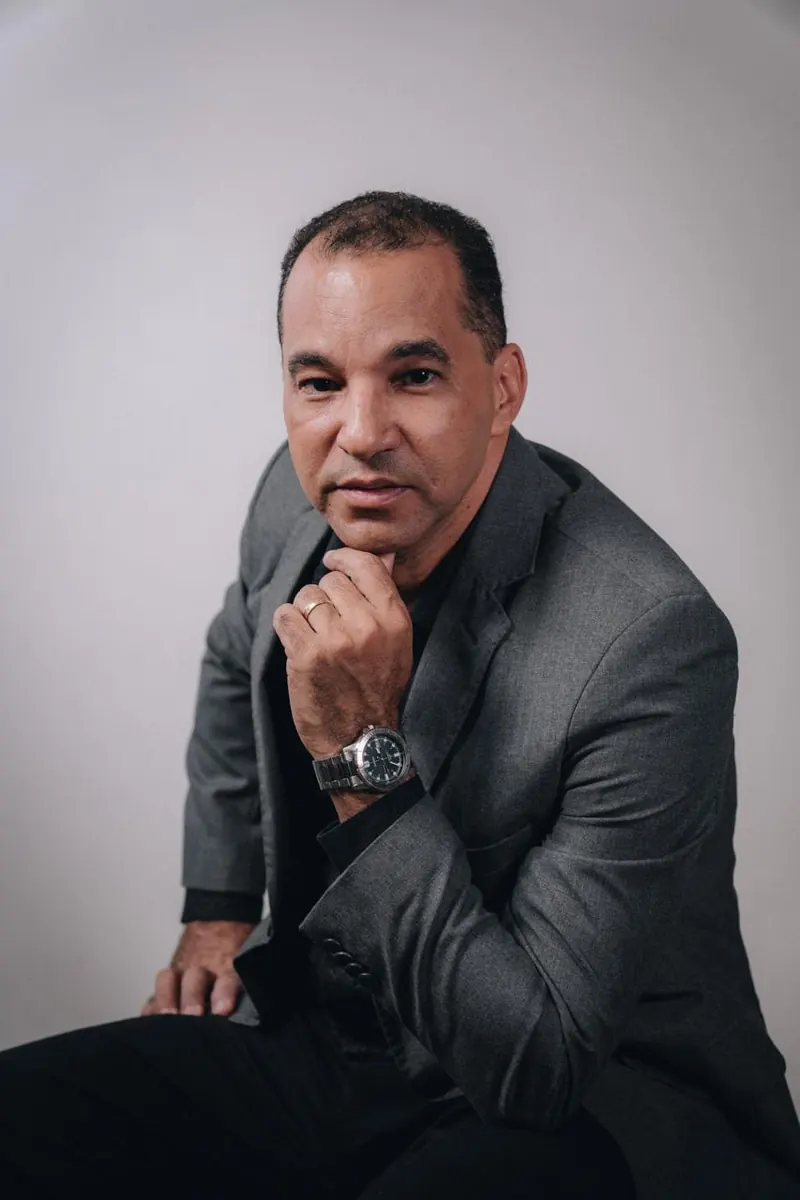 Marcos Mariano de terno cinza, com a mão no queixo, em fundo claro