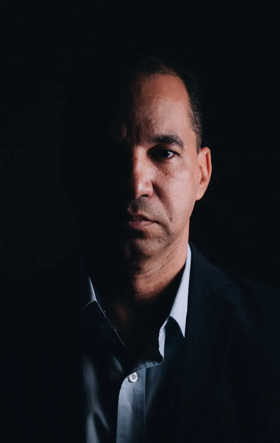 Retrato de Marcos Mariano, metade do rosto iluminada, de terno escuro